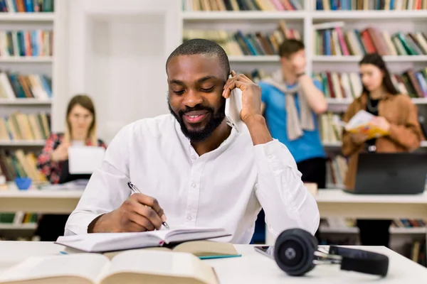 Front view of attractive smile African young man in white shirt, μιλώντας σε smartphone και γράφοντας σημειώσεις, ενώ μελετούσε και προετοιμαζόταν για εξετάσεις ή τεστ στη βιβλιοθήκη του πανεπιστημίου — Φωτογραφία Αρχείου