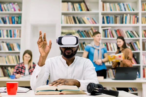Portrait d'un étudiant universitaire africain de sexe masculin lisant à la bibliothèque, portant des simulateurs de réalité virtuelle et touchant des pages de livres virtuels dans les airs. VR technologie pour l'éducation concept — Photo