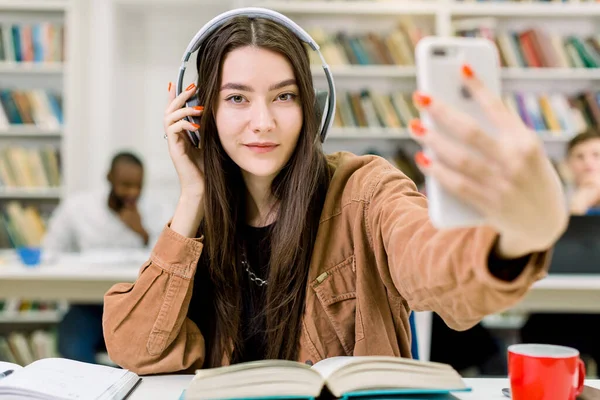 Bastante caucásica joven estudiante en ropa hipster casual, haciendo foto selfie en el teléfono inteligente para compartirlo en redes, sentado en la mesa con libros y preparándose para el examen en la biblioteca — Foto de Stock