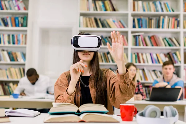 Jeune jolie fille, étudiante, utilisatrice de bibliothèque, utilisant un simulateur VR tout en faisant des recherches à la bibliothèque. Femme portant des lunettes de réalité virtuelle, assise au bureau avec de nombreux livres différents et se préparant à l'examen — Photo