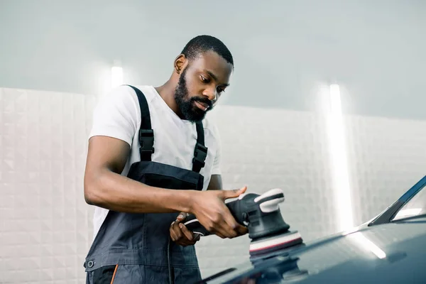 Araba temizleme servisi. Beyaz tişörtlü ve gri tulumlu yakışıklı Afrikalı Amerikalı işçi. Arabayı cilalarken orbital parlatıcısını tutuyor. Seçici odak. — Stok fotoğraf