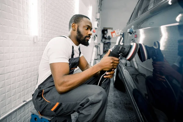 Servicio de detalle automático, pulido del coche. Vista lateral de la joven hombre afroamericano trabajador n camiseta y monos, puliendo la puerta azul del coche con pulidor orbital — Foto de Stock