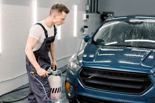 Bilservice, biltvätt och städning. Stilig ung vit manlig arbetare i t-shirt och grå overaller, gör bil förtvätt med högtrycks vattenpistol — Stockfoto