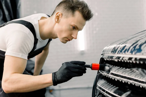 Närbild foto av ung stilig man i svart skyddshandskar rengöring radiator grill i skum med speciell borste. Biltvätt och detaljer. Tvättmaskin på stationen. Biltvätt koncept. — Stockfoto