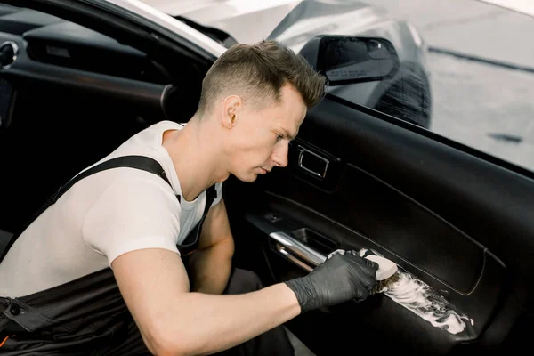 Handsome young man, worker of professional car detailing workshop, washing car door interior by a soft brush with foam. Car cleaning and detailing concept — Stock Photo, Image