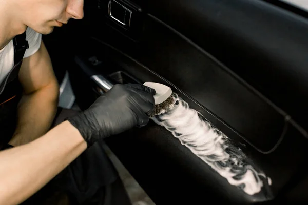 Imagem cortada de lavar o interior do carro por uma escova macia com espuma. Jovem trabalhador masculino em luvas de proteção pretas limpando a porta do carro dentro com espuma e escova — Fotografia de Stock