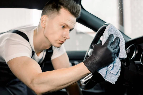 Stilig vit ung man i skyddshandskar rengöring bil interiör, bil ratt med hjälp av mikrofiber kläder. Bil detalj eller valeting koncept. Fokus på hand med tyg. — Stockfoto