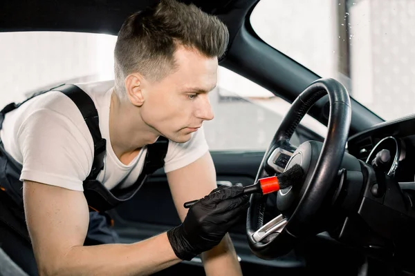 Sidovy närbild porträtt av unga kaukasiska manliga biltvätt detaljer arbetare med hjälp av rengöringsborste och ta bort damm från bilratten. Bil detalj eller valeting koncept. — Stockfoto
