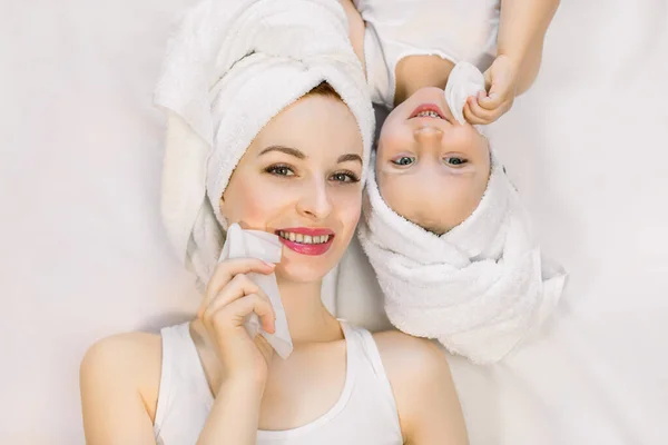Urocza młoda szczęśliwa matka i mała dwuletnia dziewczynka w ręcznikach prysznicowych na głowach, leżąca razem na białym tle i czyszcząca twarze mokrymi chusteczkami. Rodzinny dzień spa — Zdjęcie stockowe