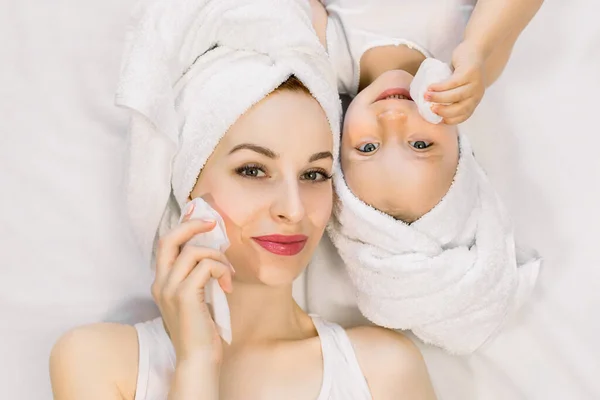 Portret czarującej młodej białej matki i jej słodkiej córeczki po kąpieli, ubranej w białe ręczniki kąpielowe na głowach, leżącej na złym i czyszczącej twarze mokrymi chusteczkami. Widok z góry — Zdjęcie stockowe