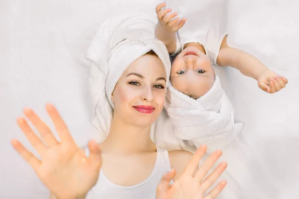 Bella sorridente giovane donna sdraiata sul letto con la sua piccola figlia carina dopo la doccia, i loro capelli sono avvolti da asciugamani bianchi freschi, stanno agitando le mani alla macchina fotografica. Famiglia spa e bagno giorno — Foto Stock