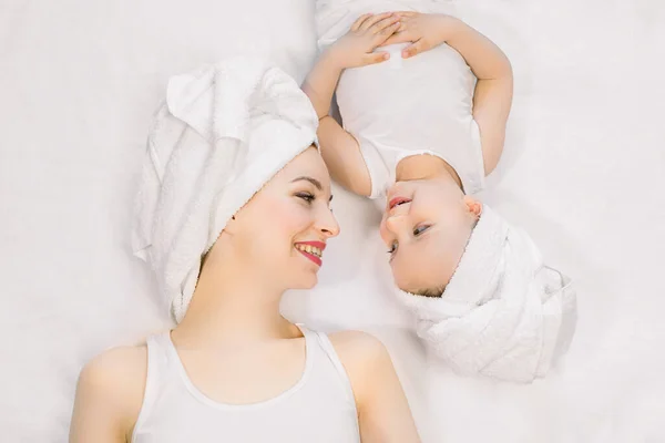 Ritratto di madre felice e figlioletta teo anni in magliette bianche e asciugamani in testa, sdraiata testa a testa sul letto e guardarsi con sorriso. Concetto di famiglia felice . — Foto Stock