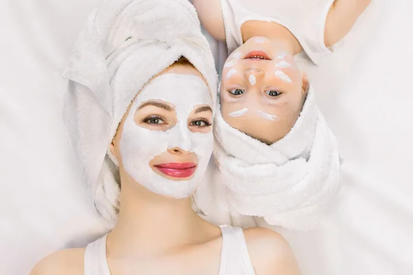 Family beauty treatment in the bathroom. Pretty mother and little two year old daughter with wrapped hair in towels, lying on white bed and making mask for a face skin. Isolated on white background. — Stock Photo, Image