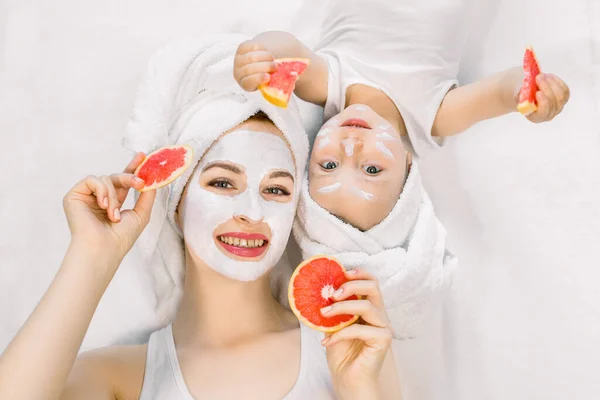 Happy family, mother and cute little daughter in white t-shirts and white towels on their heads lying on bed, doing facial beauty procedures with clay mask and holding slices of grapefruit in hands — Stock Photo, Image