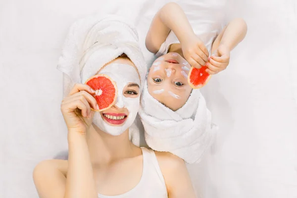 Bella giovane madre e la sua piccola figlia di due anni con asciugamani sulla testa, sdraiati insieme sul letto e facendo procedure di cura della pelle spa, maschera facciale, tenendo pezzi di pompelmo vicino ai loro occhi — Foto Stock