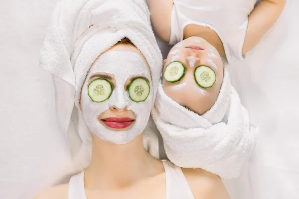 Madre e hija pequeña que se someten a procedimientos de spa juntos. Están en toallas de baño blancas en la cabeza y con rodajas de pepino en los ojos. Mujer tiene máscara facial blanca en su piel — Foto de Stock