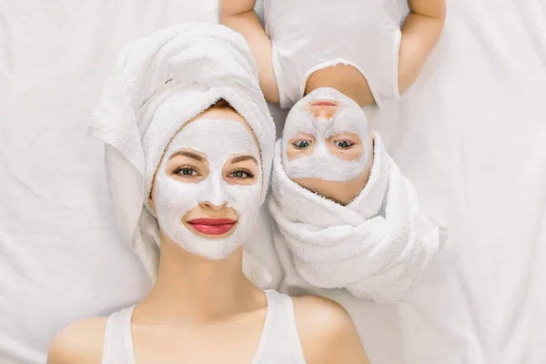 Potret ibu muda yang menyenangkan dan putri kecilnya berbaring bersama di tempat tidur dengan handuk putih di kepala dan masker tanah liat wajah sambil menikmati prosedur spa. Terisolasi, pandangan atas. Konsep kecantikan — Stok Foto