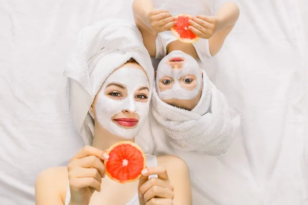 Family beauty treatment in the bathroom. Pretty mother and daughter baby girl make a clay mask for face skin, lying on bed with towels on heads and holding slices of grapefruit in hands