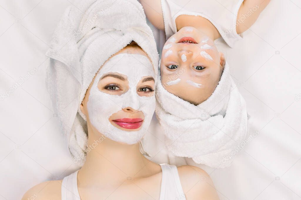 Family beauty treatment in the bathroom. Pretty mother and little two year old daughter with wrapped hair in towels, lying on white bed and making mask for a face skin. Isolated on white background.