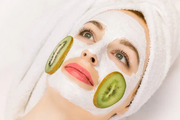 Mascarilla facial con frutas, tratamiento de belleza spa, cuidado de la piel. Chica bonita con máscara de kiwi en la cara, y rodajas de kiwi en sus mejillas, y pelo envuelto en toalla, acostado sobre fondo blanco — Foto de Stock