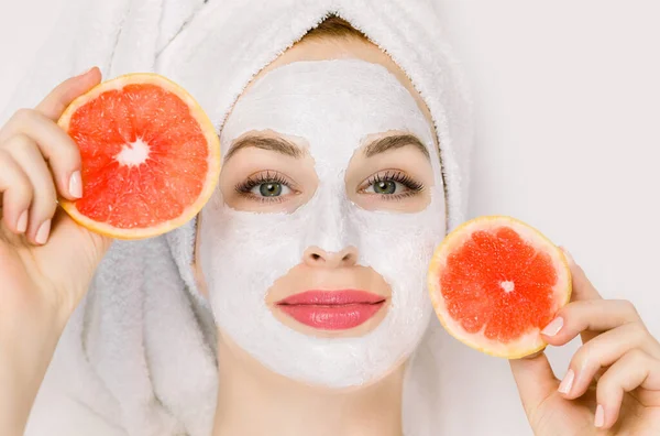 Close-up portret van mooie jonge vrouw met handdoek op het hoofd, met klei modder masker op haar gezicht, en het houden van plakjes grapefruit, liggend op witte achtergrond. Gezichtsverzorging, kosmetologie en spa — Stockfoto