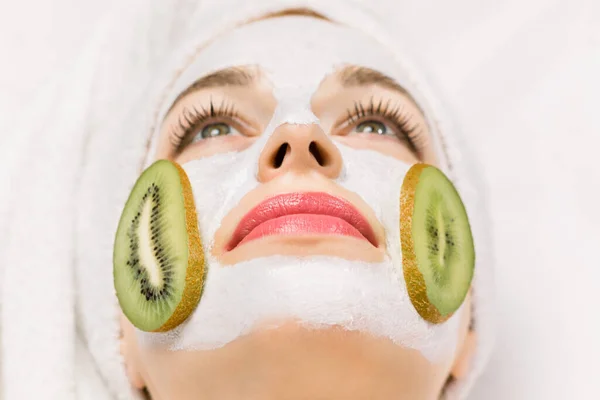 Skin care, facial mask and cosmetology concept. Close up macro of face of pretty woman with mud facial mask and kiwi fruit slices on her cheeks, lying on white background.