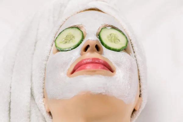 Close-up beeld van jonge vrouw met haar gewikkeld in badhanddoek, met klei gezichtsmasker en komkommer plakjes op haar ogen, liggend op geïsoleerde witte achtergrond. Bovenaanzicht, macro, kopieerruimte — Stockfoto