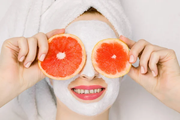 Mooi jong meisje in badhanddoek en gezichtsmasker met grapefruit plakjes voor haar ogen. mooie vrouw houden grapefruit plakjes als bril, verbergen haar ogen, liggend op witte achtergrond — Stockfoto