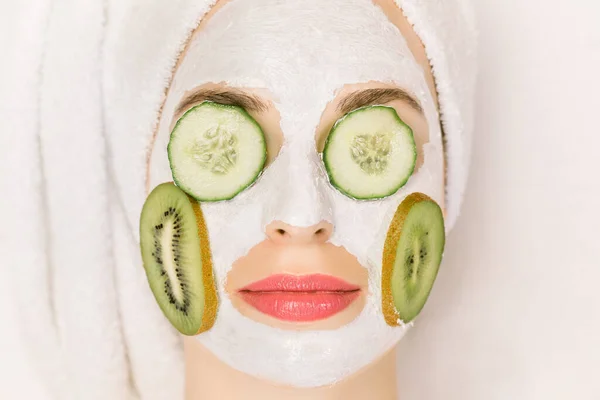 Konsep perawatan kulit dan kosmetologi. Top view, close up of woman with clay mud mask on her face, with slices of kiwi on cheeks and pieces of cucumber cover eyes, lying on white background — Stok Foto