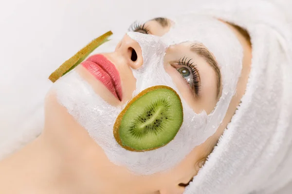 Tratamiento facial, cuidado de la piel, spa en casa. Foto de vista lateral de la joven en toalla en la cabeza, con mascarilla facial de fruta casera natural en la piel y rodajas de kiwi fresco en las mejillas . — Foto de Stock