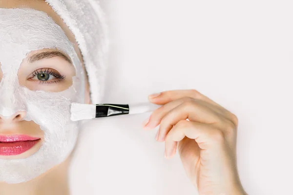 Gambar wanita muda cantik dan menarik dengan rambut yang dibungkus handuk mandi putih, mengenakan masker lumpur dengan kuas, berbaring di latar belakang putih yang terisolasi. Profesional wajah perawatan kecantikan kulit — Stok Foto