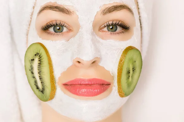 Prosedur spa dan kosmetologi. Tutup bagian atas wajah wanita muda yang menarik dengan topeng buah di wajahnya wanita dan irisan kiwi di pipi, dan dengan handuk mandi di kepala di spa saloon — Stok Foto