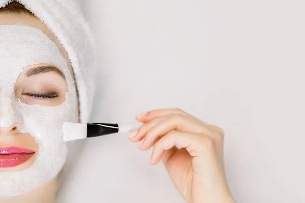 Φροντίδα δέρματος. Κοντινό πλάνο της νεαρής γυναίκας με πετσέτα στο κεφάλι και με θρεπτική μάσκα προσώπου στο πρόσωπο, χαλαρώνοντας στο σαλόνι ομορφιάς με τα μάτια κλειστά, κρατώντας πινέλο για την εφαρμογή της μάσκας. Πάνω όψη, αντιγραφή χώρου — Φωτογραφία Αρχείου