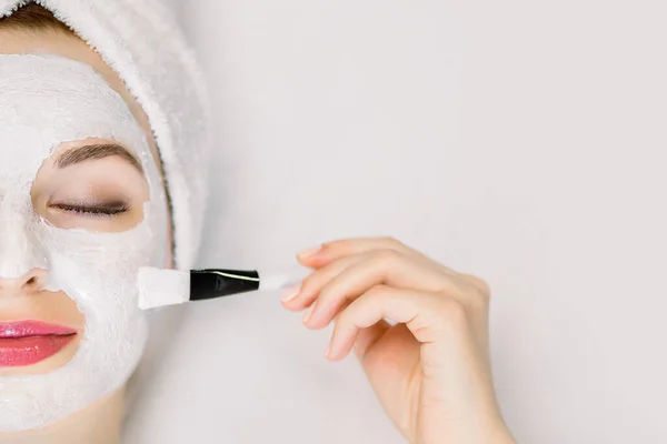 Cuidado com a pele. Close up de jovem com toalha na cabeça e com máscara facial nutriente no rosto, relaxando no salão de beleza com os olhos fechados, segurando escova para aplicação de máscara. Vista superior, espaço de cópia — Fotografia de Stock