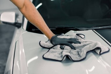 Araba temizliği, yıkama ve temizlik konsepti. Siyah lastik eldivenli, gri mikrofiberi tutan ve lüks beyaz arabanın kaputunu cilalayan profesyonel erkek araba yıkama işçisinin kırpılmış görüntüsü.