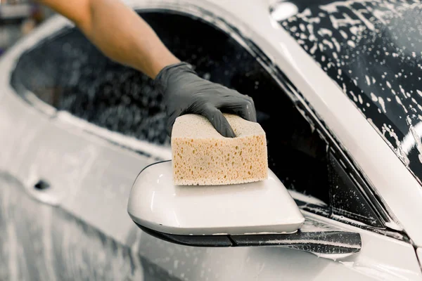 Imagem cortada da mão do trabalhador de lavagem de carro lavando um espelho ensaboado de carro de luxo branco, segurando esponja. Concentre-se na mão em luva preta, segurando esponja e lavando espelho do carro. Limpeza, conceito de lavagem de carro — Fotografia de Stock