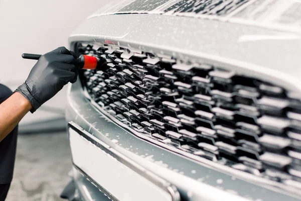 Lavado de detalles del coche. Imagen recortada de la mano del trabajador masculino en guantes de goma lavando la rejilla del radiador del coche de lujo coche blanco con cepillo especial — Foto de Stock