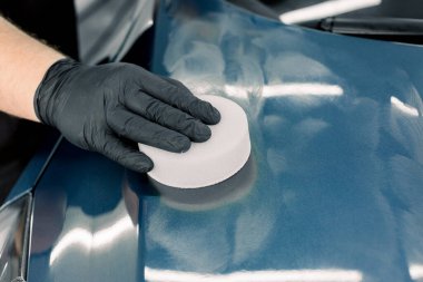 Erkek elinin koruyucu siyah eldivenli yakın plan görüntüsü, sağlam carnauba cilasıyla beyaz sünger tutmak, ve profesyonel temizlik atölyesinde lüks mavi arabanın kaputunu cilalamak. Araba temizleme