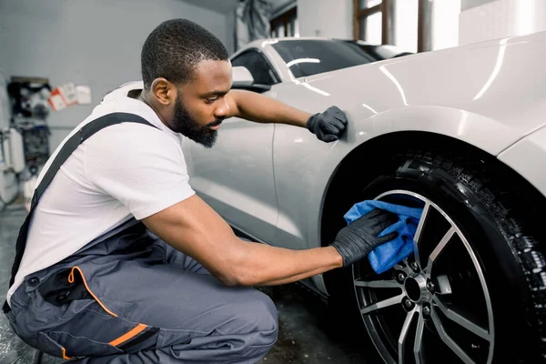 Lavagem de carro e detalhando foto. Africano homem trabalhador em macacões de proteção e luvas de borracha, lavar roda liga de carro em uma lavagem de carro, usando pano de microfibra e limpador especial — Fotografia de Stock