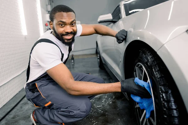 Araba yıkama servisinde elle yıkama. Yakışıklı koyu tenli erkek işçi modern beyaz arabanın tekerlek kenarını mavi mikrofiber kumaşla temizliyor, kameraya bakıyor. — Stok fotoğraf