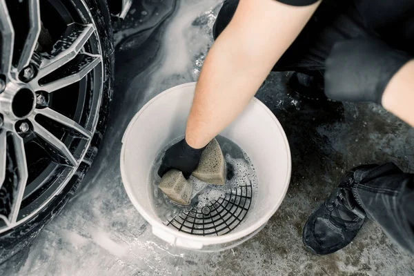 Lavagem de carro e detalhando conceito. Vista superior de ferramentas de lavagem no serviço de lavagem de carro, balde branco com solução de limpeza de sabão, grade especial. Mão de trabalhador masculino segurando esponja para limpar jantes de carro — Fotografia de Stock