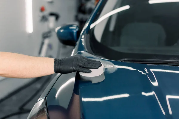 Cortada imagem de perto da mão masculina em luva preta protetora, segurando esponja branca com cera de carnaúba sólida e capa de polimento de carro azul de luxo na oficina de detalhamento profissional. Detalhes do carro — Fotografia de Stock