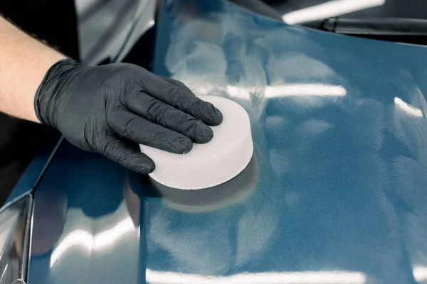 Beskuren närbild av manlig hand i skyddande svart handske, håller vit svamp med fast karnaubavax, och polering huva av lyx blå bil på professionell detaljering verkstad. Detaljerad bilbeskrivning — Stockfoto