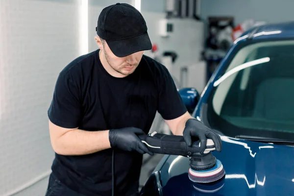 Bildetaljer och polering koncept. Professionell vit manlig bil servicearbetare, bär svart t-shirt och mössa, hålla i händerna omloppsbana polerare, och polering blå lyx bil i bilverkstad — Stockfoto