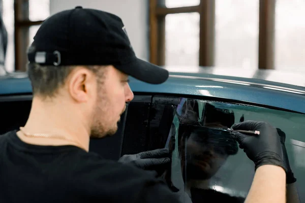 Vista posterior del ángulo del coche que detalla al trabajador masculino del taller, usando la gorra negra y la camiseta, aplicando la lámina del tinte en una ventana del coche en un garaje — Foto de Stock