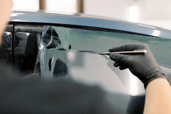 Imagem cortada das mãos do trabalhador na garagem pintando uma janela de carro com folha ou filme colorido, segurando lâmina ou faca especial para cortar o filme. Oficina de detalhamento de carro, vidros de tintura — Fotografia de Stock