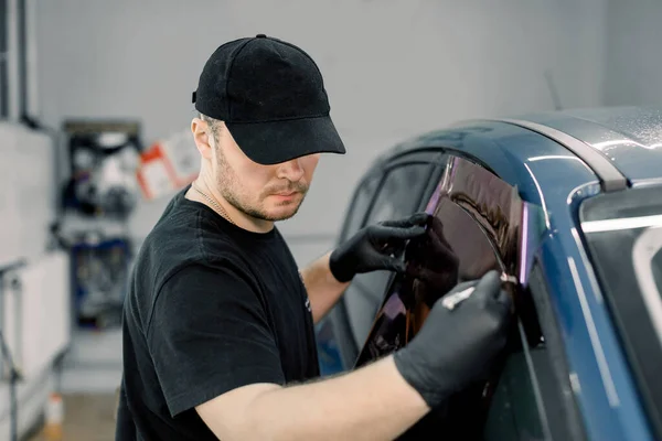 Imagen de primer plano de un trabajador mecánico de automóviles guapo, con uniforme negro, adjuntando lámina de teñido a la ventana del coche en la estación de servicio especializada —  Fotos de Stock