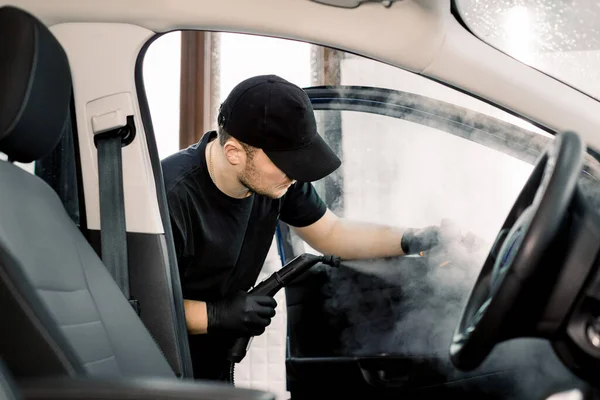Oto temizleme ve detay verme servisi. Siyah üniformalı yakışıklı beyaz adam arabanın içini sıcak buhar temizleyicisiyle temizliyor. Seçici odak. — Stok fotoğraf