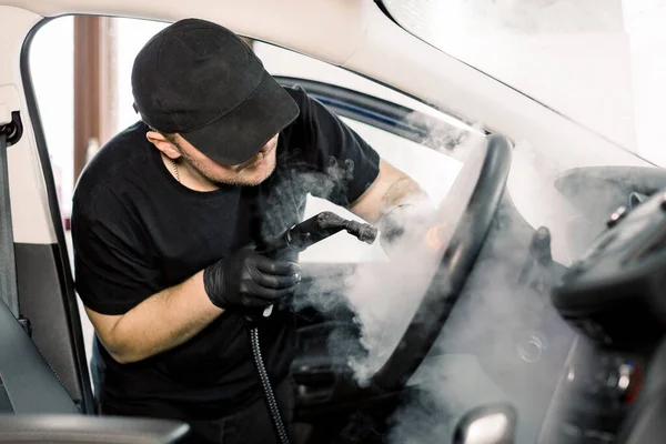 Araba temizliği, buhar temizleme konsepti. Siyah tişörtlü ve şapkalı yakışıklı bir adam. Araba yıkama merkezinde çalışıyor. İçini sıcak buharlı temizleyiciyle temizliyor. Araba temizleme kavramı Stok Resim