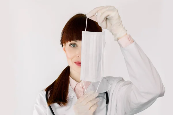 Молода красива темноволоса жінка-лікар або медсестра, одягнена в біле пальто, ховає обличчя за медичною маскою, позує на ізольованому білому тлі, копіює простір — стокове фото